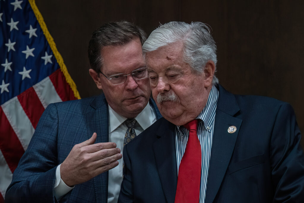A man wearing a suit and glasses whispers into the ear of a much older man wearing a suit.